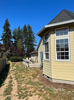 Sunny yellow house with large windows and wooden deck, surrounded by tall trees and clear blue sky.