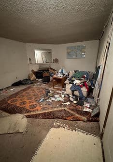 Cluttered living room with trash, disorganized furniture, and a stained rug.